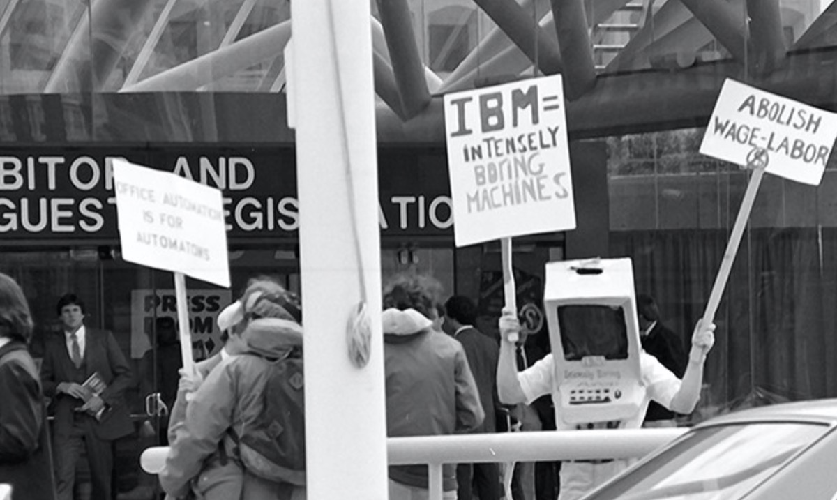 Image of people in front of the entrance to a convention center, one wearing a computer costume and holding a placard that reads IMB = intensely boring machines. Image of people in front of the entrance to a convention center, one wearing a computer costume and holding a placard that reads IMB = intensely boring machines.