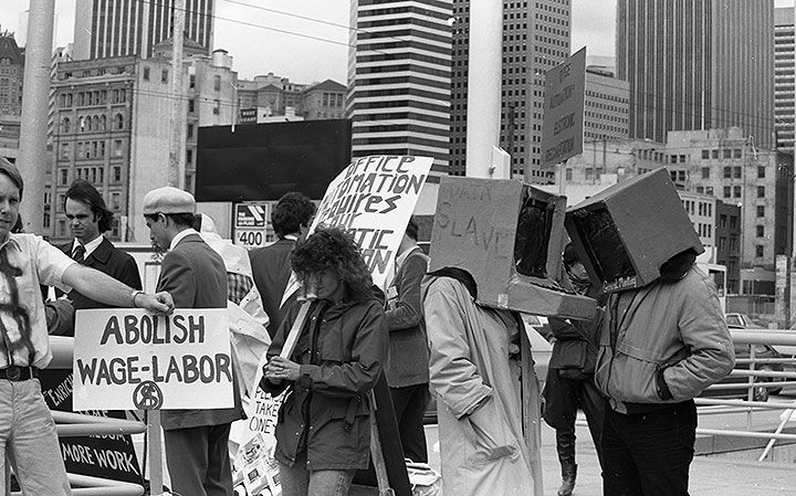 Image of people in front of the entrance to a convention center with skyscrapers in the background and holding placards that read ABOLISH WAGE-LABOR AND OFFICE AUTOMATION REQUIRES OUR... two people are wearing computer costumes on their head, one with the word DATA SLAVE written on the side. Image of people in front of the entrance to a convention center with skyscrapers in the background and holding placards that read ABOLISH WAGE-LABOR AND OFFICE AUTOMATION REQUIRES OUR... two people are wearing computer costumes on their head, one with the word DATA SLAVE written on the side.