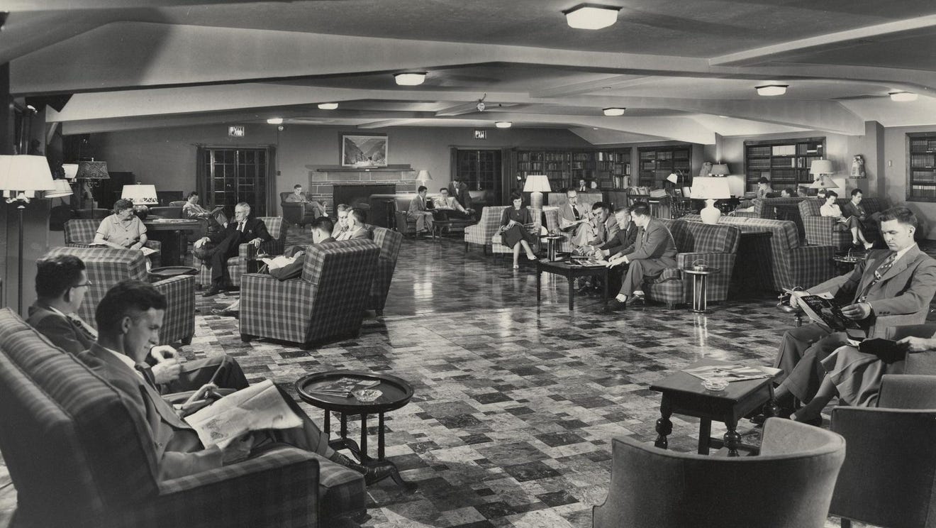 Black-and-white photograph of a large, fancy-looking room with about a dozen people sitting in sofas, reading newspapers and talking. Most of the people depicted are men wearing suits, though there are a few women present. Black-and-white photograph of a large, fancy-looking room with about a dozen people sitting in sofas, reading newspapers and talking. Most of the people depicted are men wearing suits, though there are a few women present.