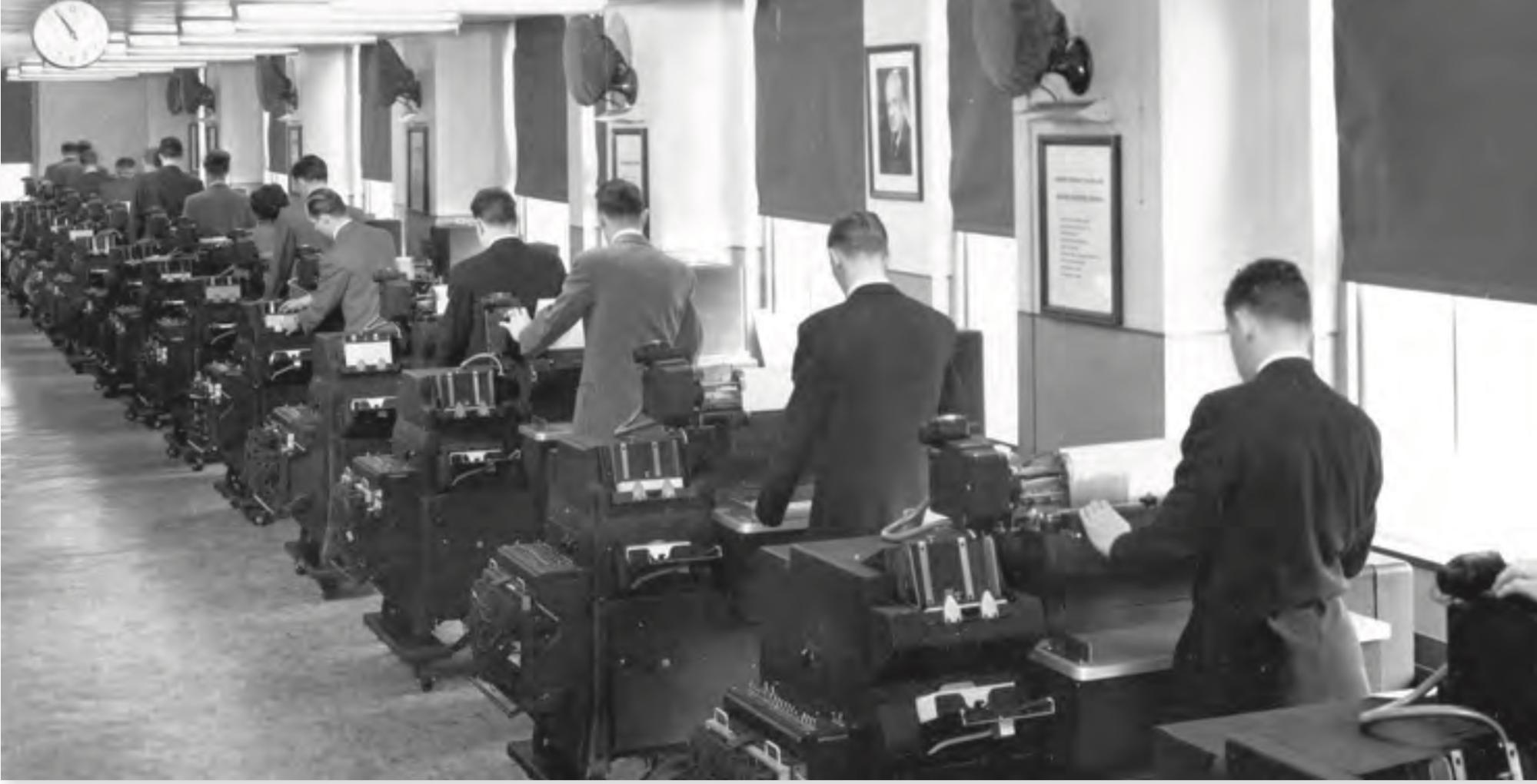Black-and-white photograph depicting a row of suit-wearing men standing in front of their desks, operating old computer equipment. Black-and-white photograph depicting a row of suit-wearing men standing in front of their desks, operating old computer equipment.