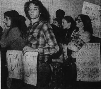 Black-and-white photograph of protesters carrying anti-apartheid signs, including a bespectacled man holding up a sign that says 'DIVEST' Black-and-white photograph of protesters carrying anti-apartheid signs, including a bespectacled man holding up a sign that says 'DIVEST'