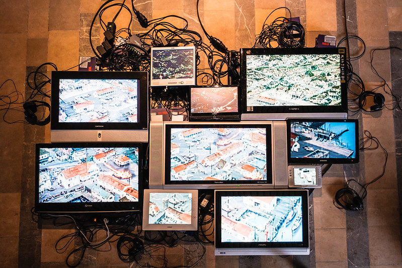 Several screens are assembled into an art installation, showing images of buildings and piers, with tangled wires visible in the background. Several screens are assembled into an art installation, showing images of buildings and piers, with tangled wires visible in the background.