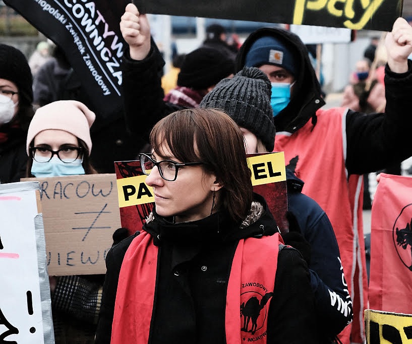 Magda, with glasses and a red vest with a wildcat cat icon on it, stands with fellow Amazon workers and Inicjatywa Pracownicza union members Magda, with glasses and a red vest with a wildcat cat icon on it, stands with fellow Amazon workers and Inicjatywa Pracownicza union members