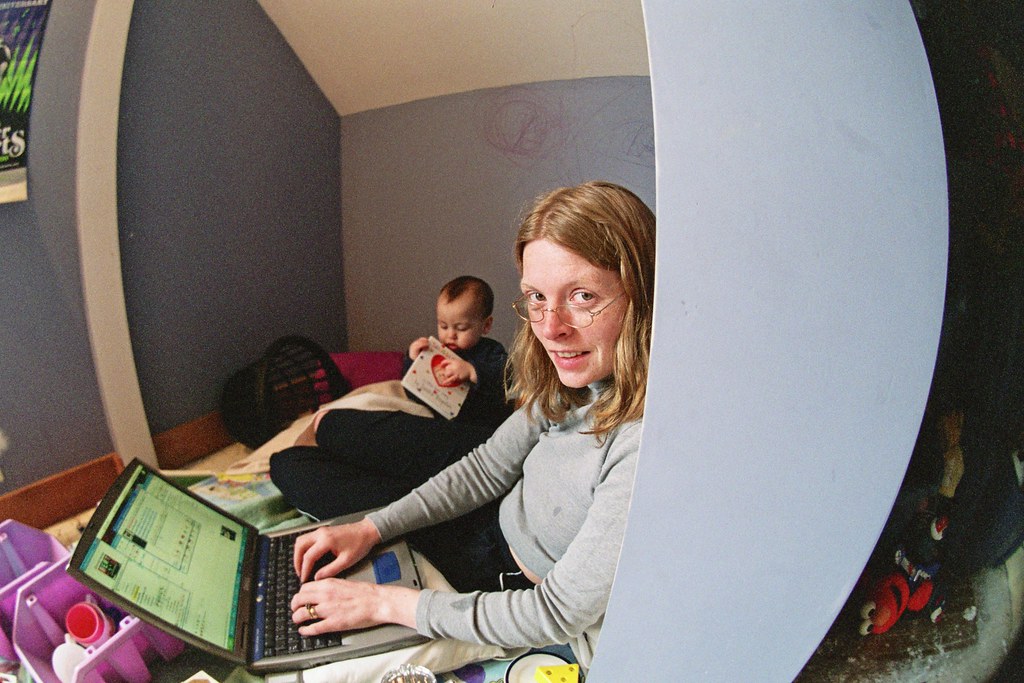 A woman working on her computer with a toddler nearby. A woman working on her computer with a toddler nearby.