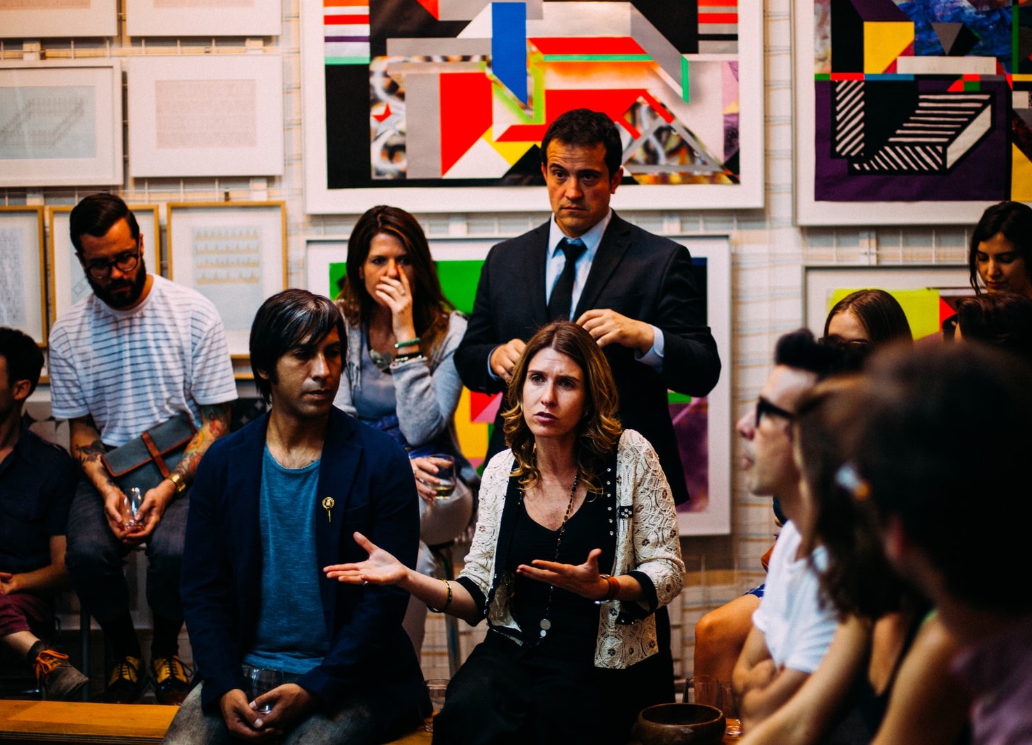 A person with long hair and gesturing with hands out appears to share a story in a room with a dozen other people in business casual attire and modern art on the wall behind them A person with long hair and gesturing with hands out appears to share a story in a room with a dozen other people in business casual attire and modern art on the wall behind them