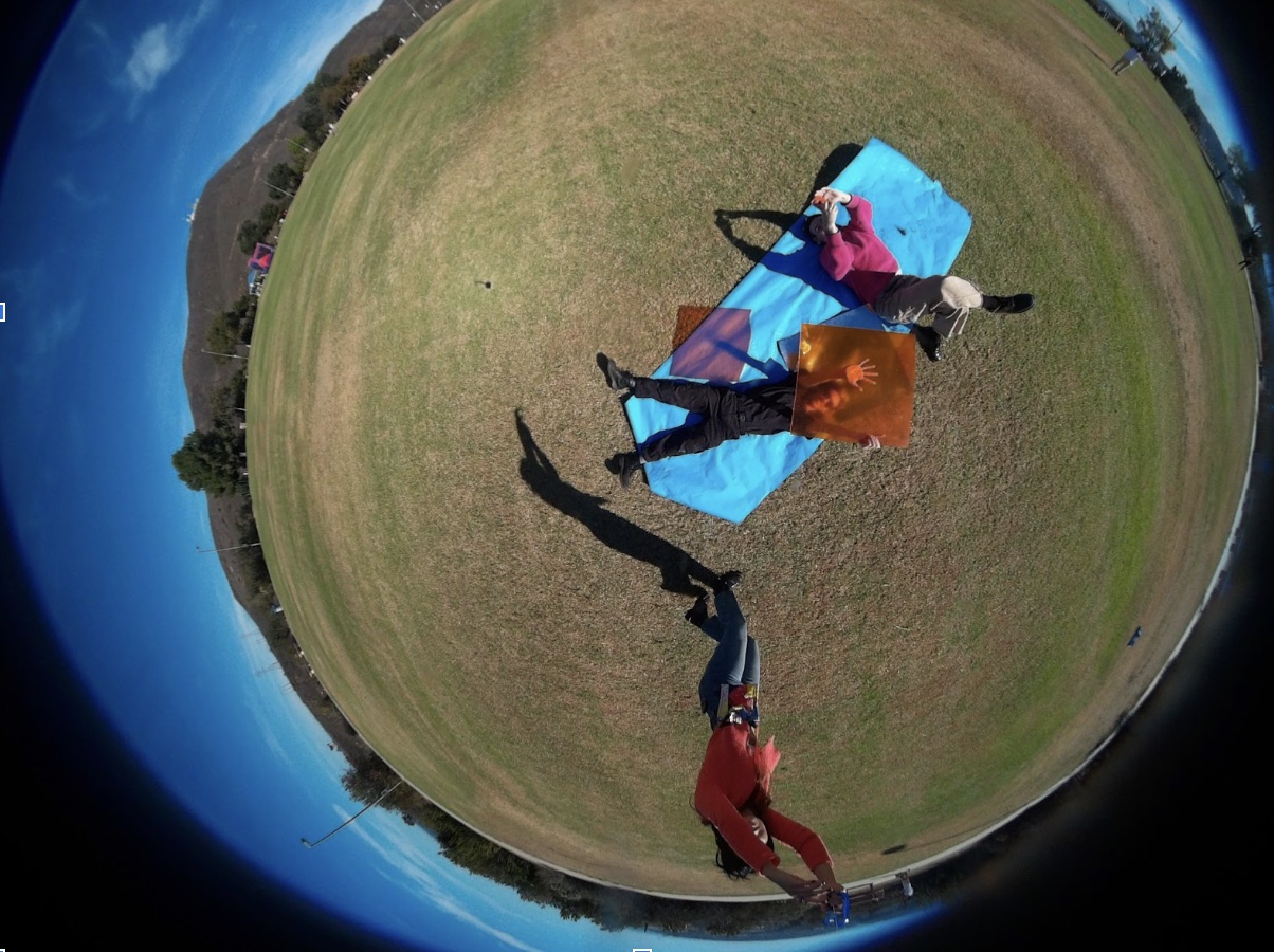 One person holds the string of the kite, while another holds an orange filter over their face, and a third takes the photograph using a phone app connected to the kite camera. One person holds the string of the kite, while another holds an orange filter over their face, and a third takes the photograph using a phone app connected to the kite camera.
