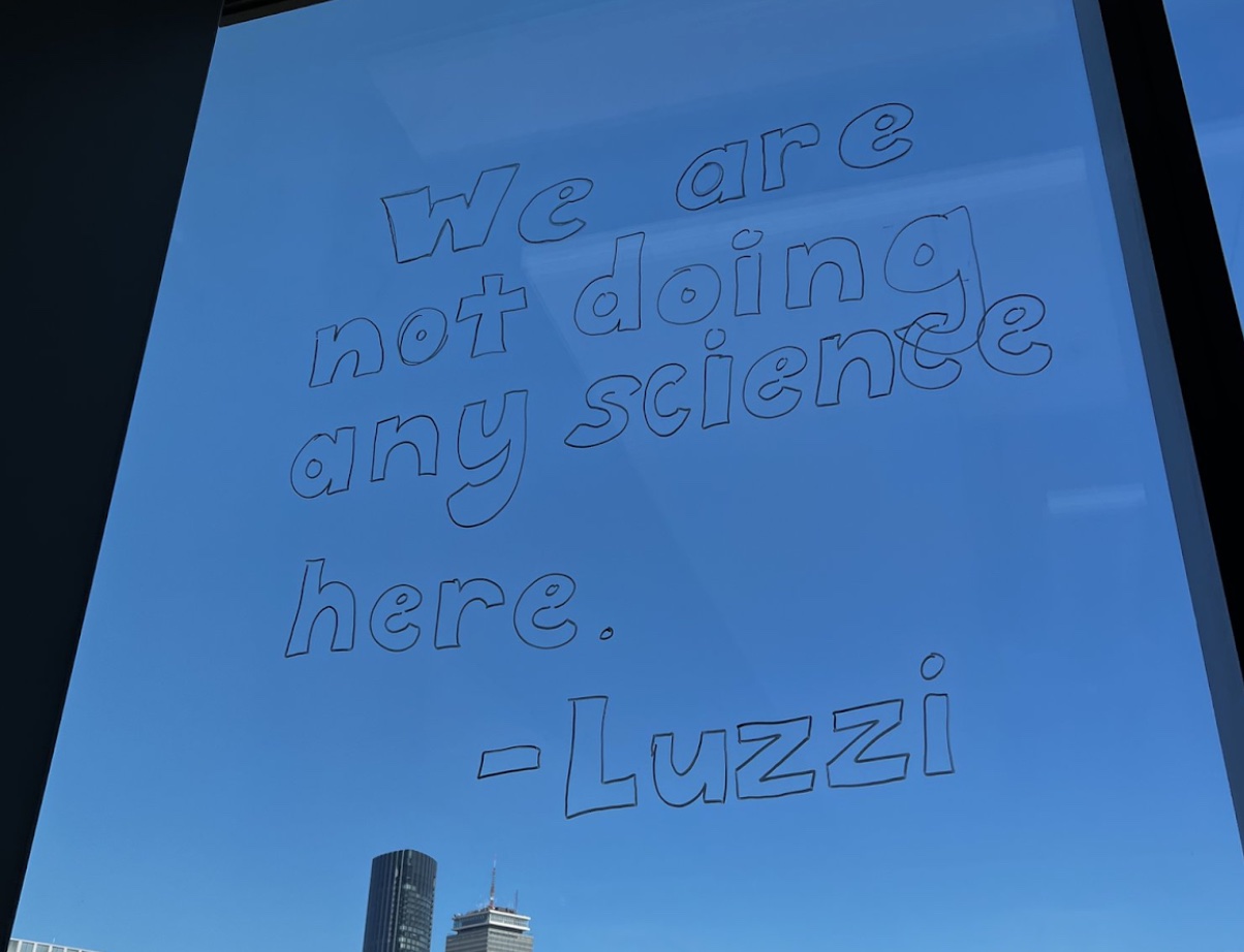 A window looking out onto the Boston skyline and a clear blue sky with writing in dry-erase marker that reads, WE ARE NOT DOING ANY SCIENCE HERE - LUZZI A window looking out onto the Boston skyline and a clear blue sky with writing in dry-erase marker that reads, WE ARE NOT DOING ANY SCIENCE HERE - LUZZI