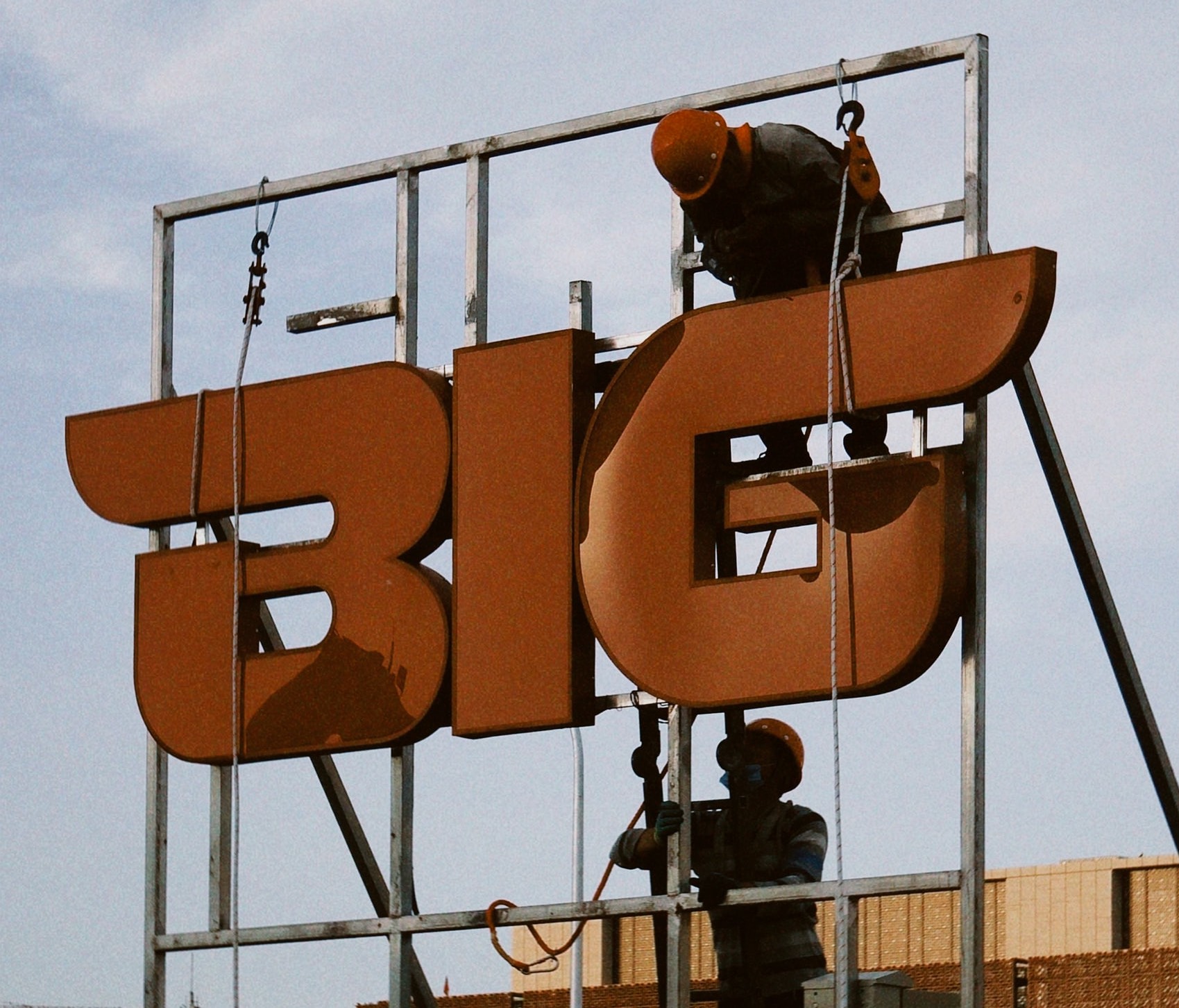 Two workers on the job, talking it out as they install an organgle rooftop sign that says BIG Two workers on the job, talking it out as they install an organgle rooftop sign that says BIG