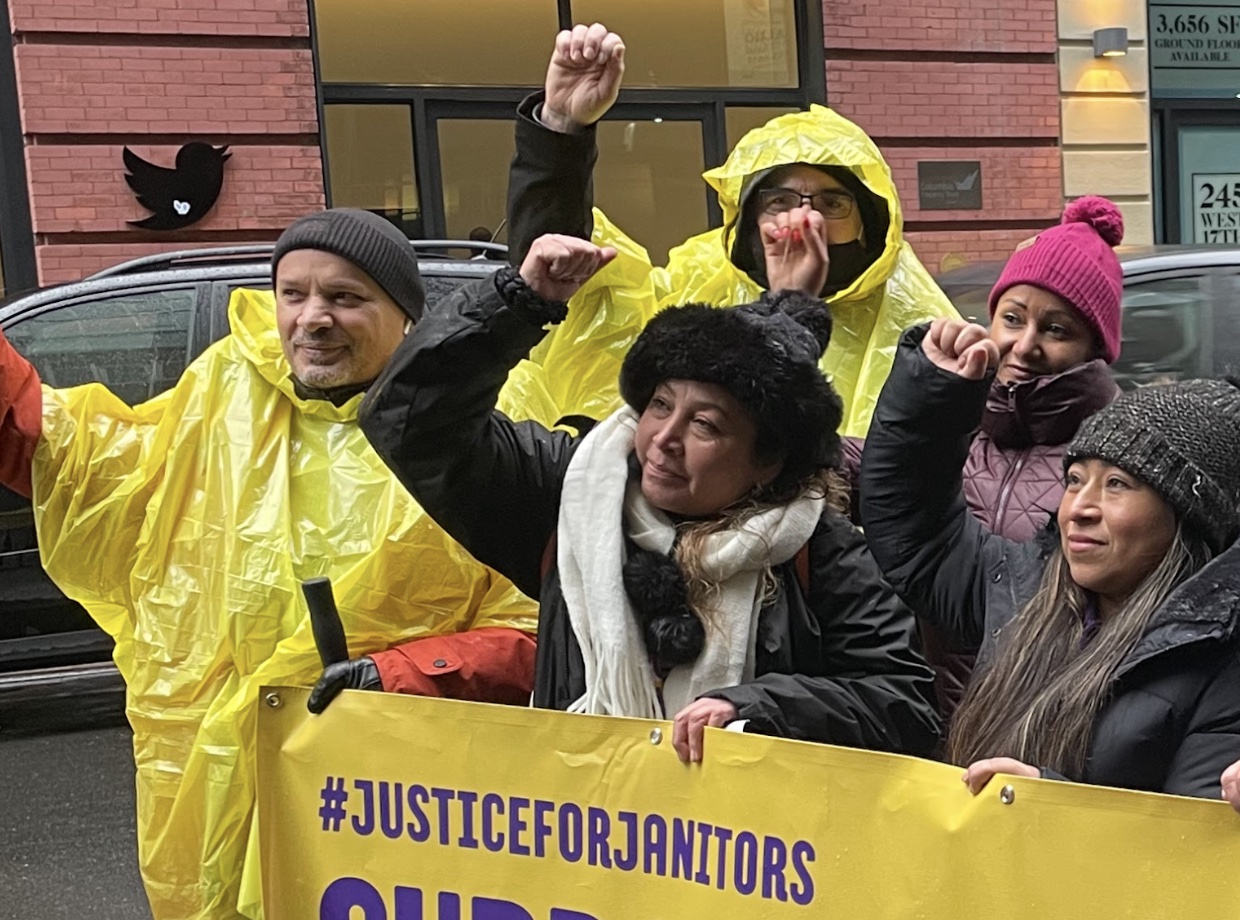 A group of workers in yellow rain jackets holing a banner in one hand that says hashtag justice for janitors and all raising their other fist high, standing in front of the NYC Twitter building and its logo on the wall in the background A group of workers in yellow rain jackets holing a banner in one hand that says hashtag justice for janitors and all raising their other fist high, standing in front of the NYC Twitter building and its logo on the wall in the background